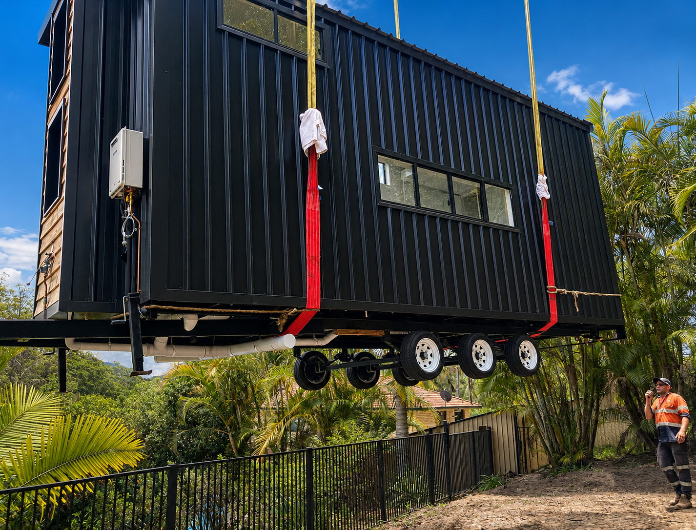 A completed tiny home being lifted by crane straps into place, with a tradesperson on site