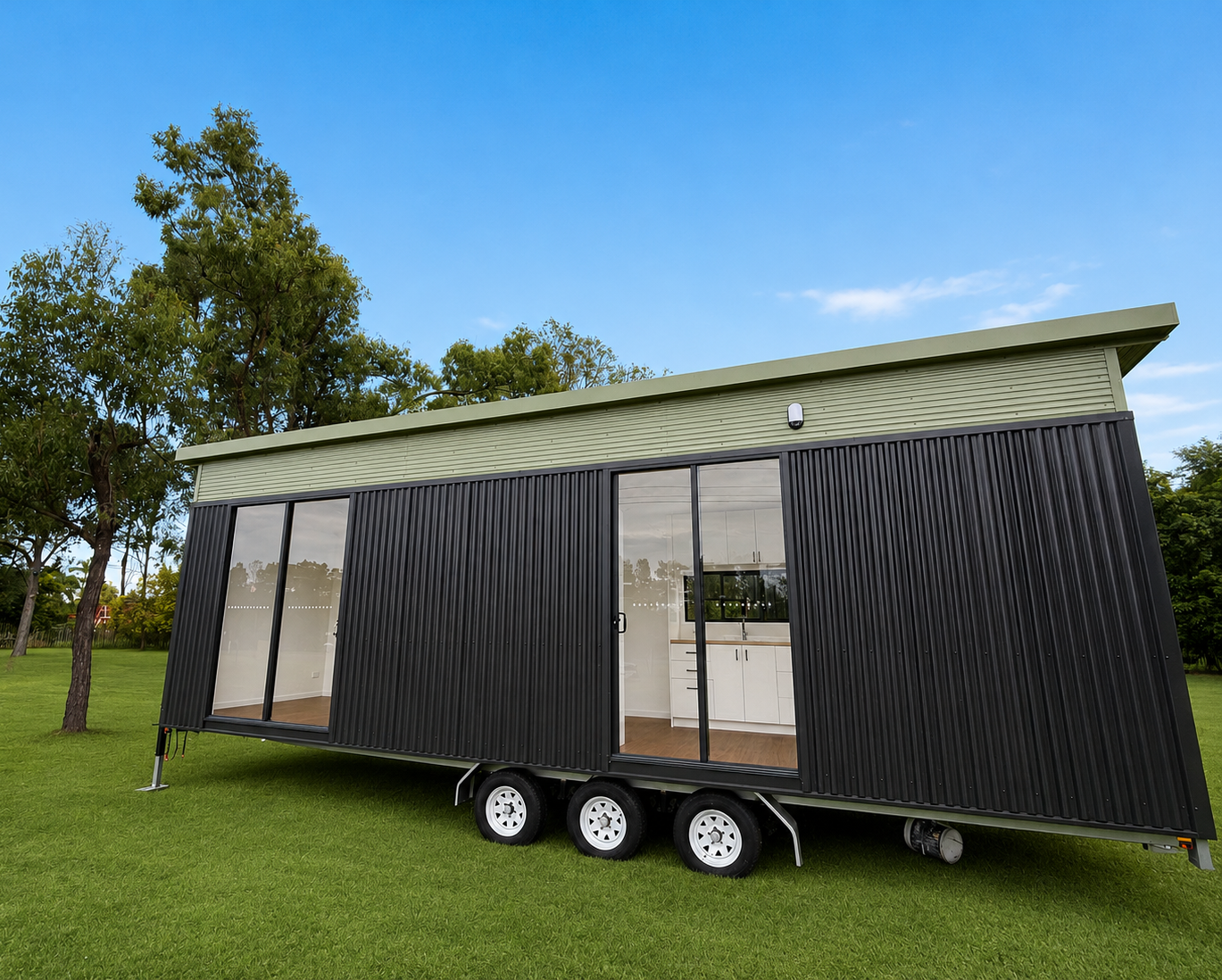 A modern black tiny home with an olive-green roofline and full-height glass sliders, on a registered trailer on green lawn under a blue Queensland sky
