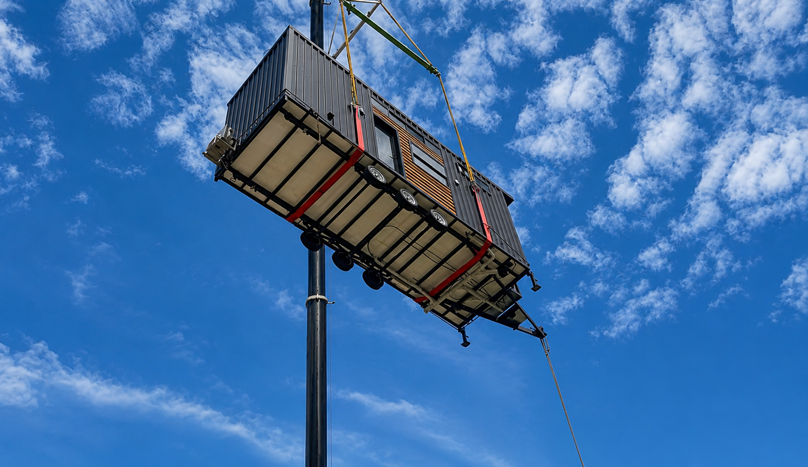 A finished tiny home lifted by crane against a blue cloud-streaked sky during delivery