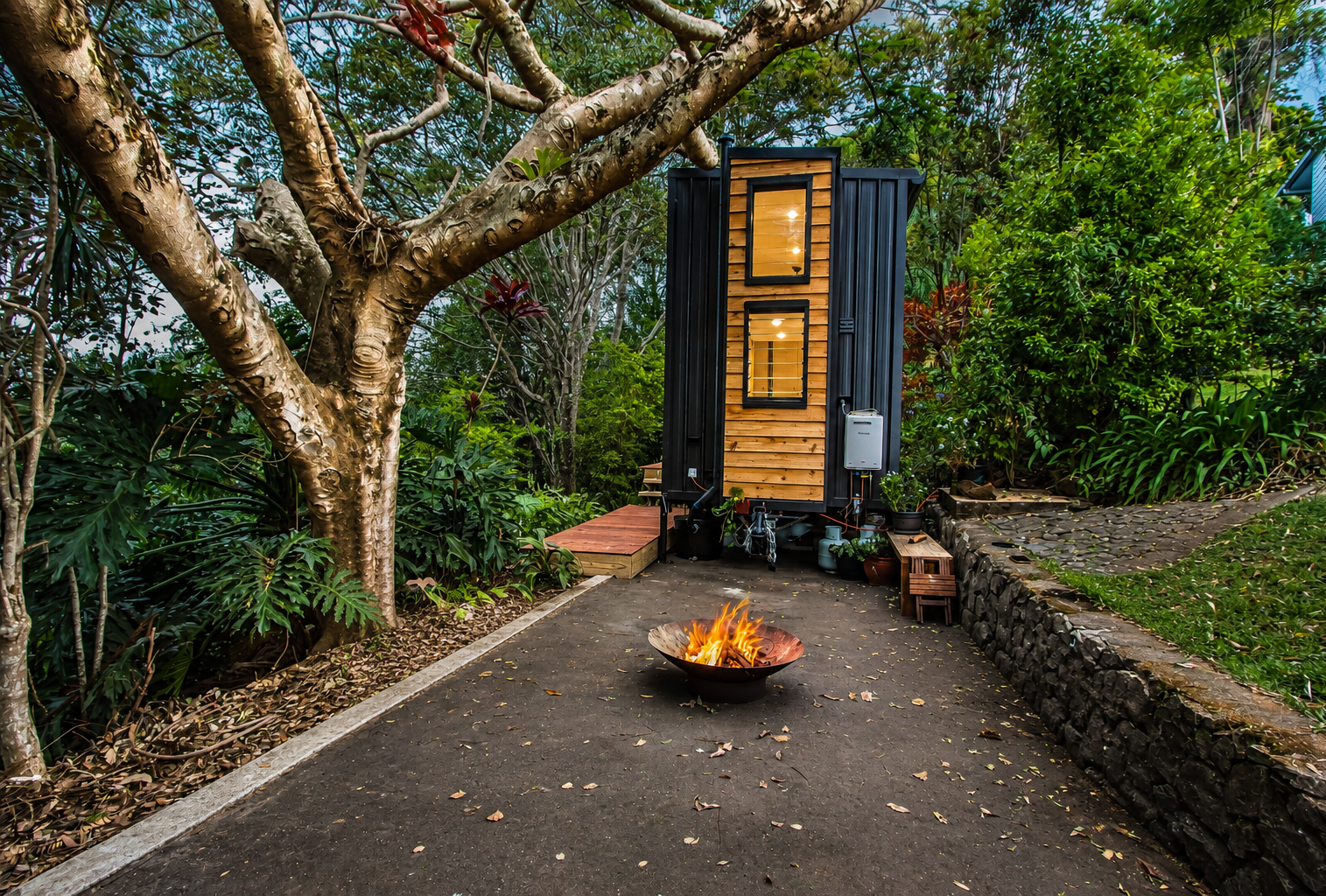 Two-storey timber and black tiny home in a lush garden with a fire bowl