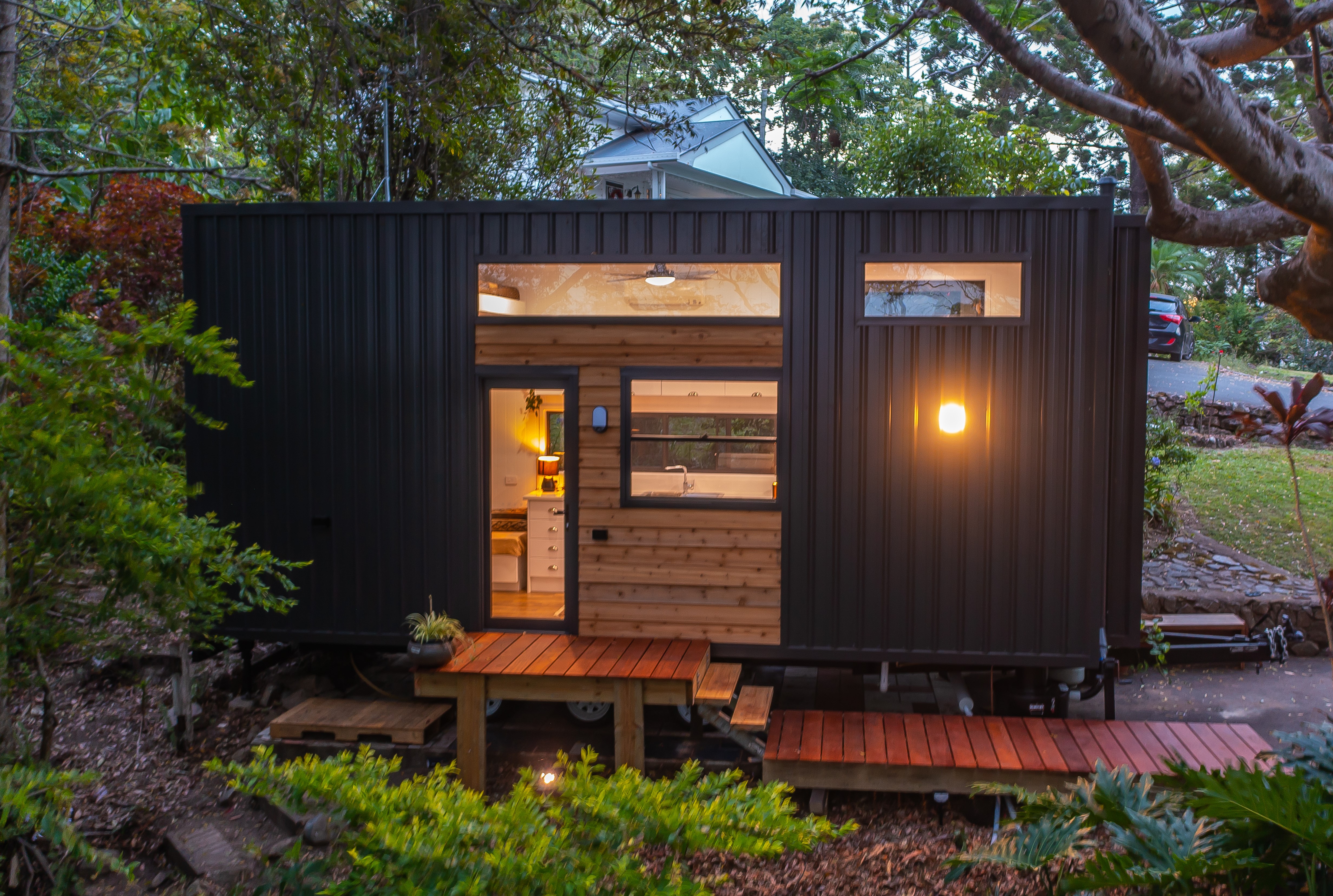A completed tiny home at dusk with warm lit windows in a leafy garden